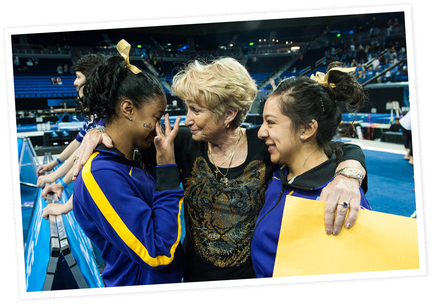 Three women embracing in a sports arena, with two wearing blue and yellow athletic jackets, and the middle woman with short blonde hair and a black shirt with a tiger design, sharing an emotional moment.