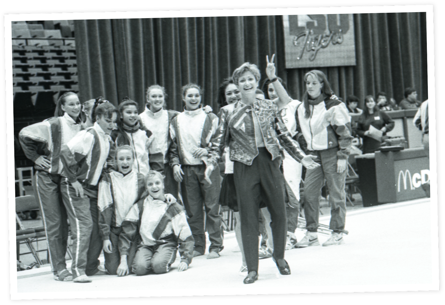 D-D in a patterned blazer and pants leading a group of young girls in track suits, in a gymnasium with a curtain in the background.