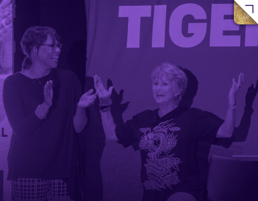Two women smiling and high-fiving in front of a purple-lit background with the word "TIGER" partially visible.