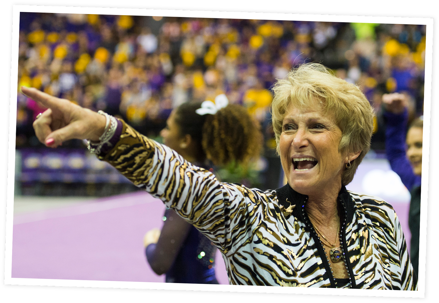 D-D Breaux with short blonde hair, wearing a zebra print jacket and jewelry, is pointing and shouting towards a sports event with a crowd of spectators in the background.
